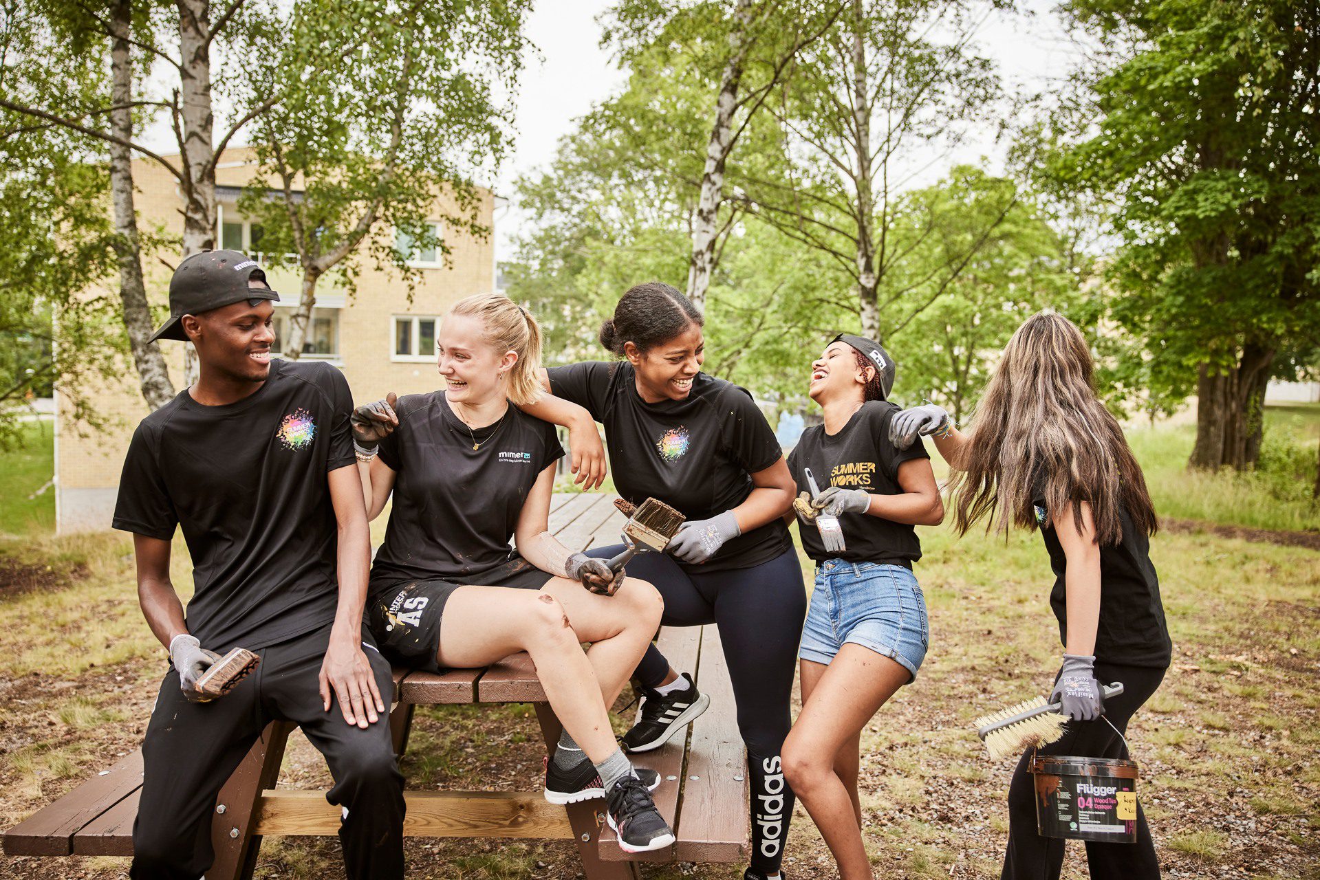 5 ungdomar i SummerWorks-kläder skrattar på parkbänk.
