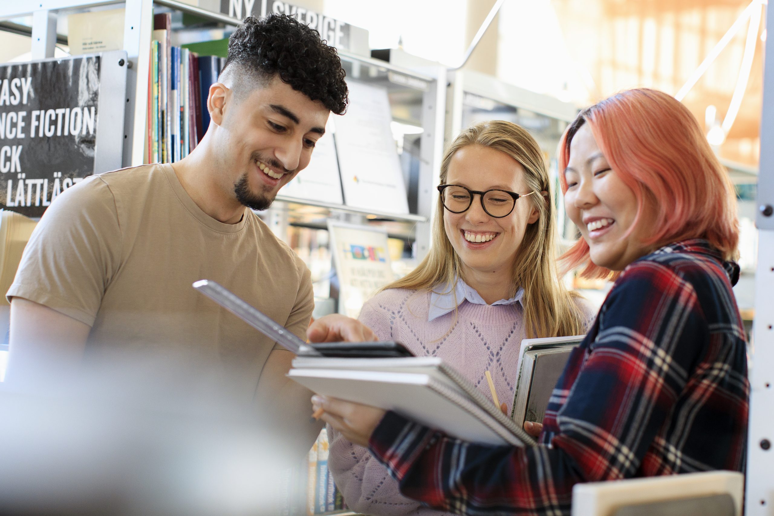 En kille och tre tjejer står och ler på ett bibliotek. De tittar på en bok.